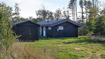 A two-story cabin with a front door and a small window on the upper floor, surrounded by a grassy area and tall trees.