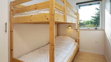 A bunk bed with a wooden frame, a single bed below, a window with a view of greenery, and a wall-mounted light fixture.