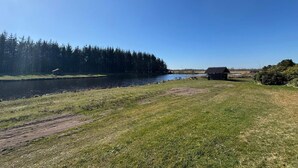 A lakeside view with a small cabin, picnic tables, and a bird flying near the water. - 5 Star Holiday Home in Ringkobing (Ringkobing)