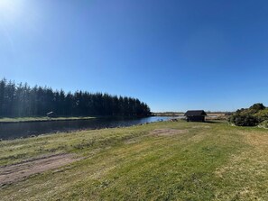 A lakeside view with a small cabin, picnic tables, and a bird flying near the water. - 5 Star Holiday Home in Ringkobing (Ringkobing)