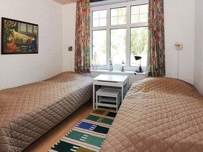 A room with two beds, a small desk, a window with curtains, a painting on the wall, and a rug on the floor.