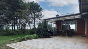 Une terrasse en bois équipée d’un barbecue, de meubles d’extérieur et d’une véranda en verre.
