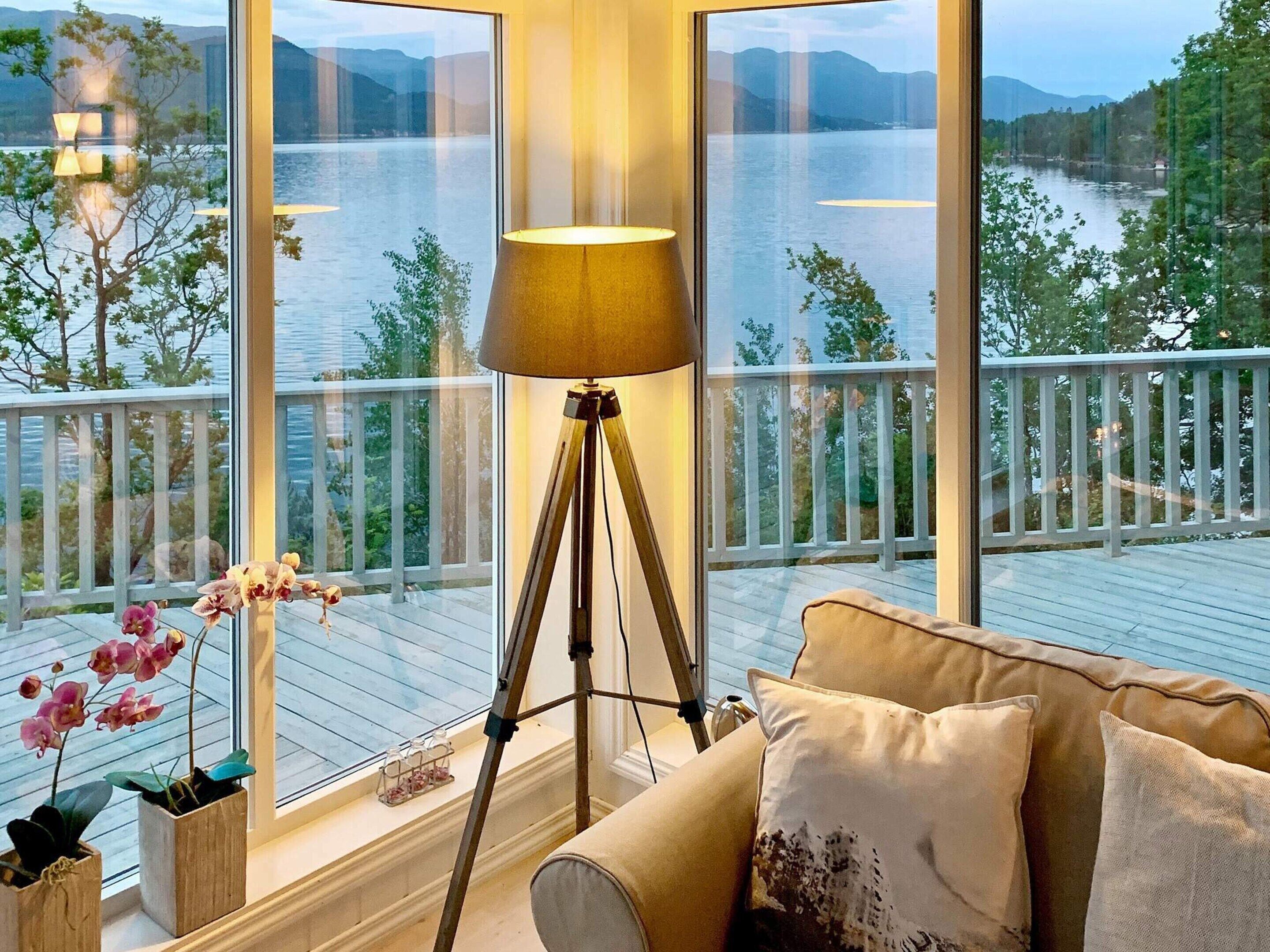 A room with a large window overlooking a lake, a floor lamp, and a sofa with cushions.