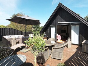 Een houten terras met rieten meubilair, een grote parasol en de ingang van een gebouw.