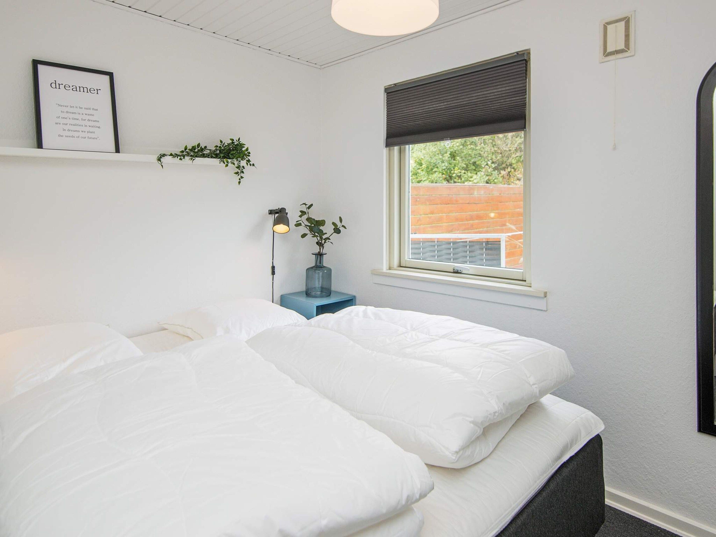 Ein Schlafzimmer mit einem Bett, einem Nachttisch mit Lampe, einem Fenster mit Blick und einem gerahmten Zitat an der Wand.