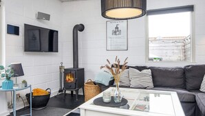 A living room with a white sofa, a glass coffee table, a black wood-burning stove, a TV mounted on the wall, and a window with a view of a building. - 4 Star Holiday Home in Hejls (Hejls)