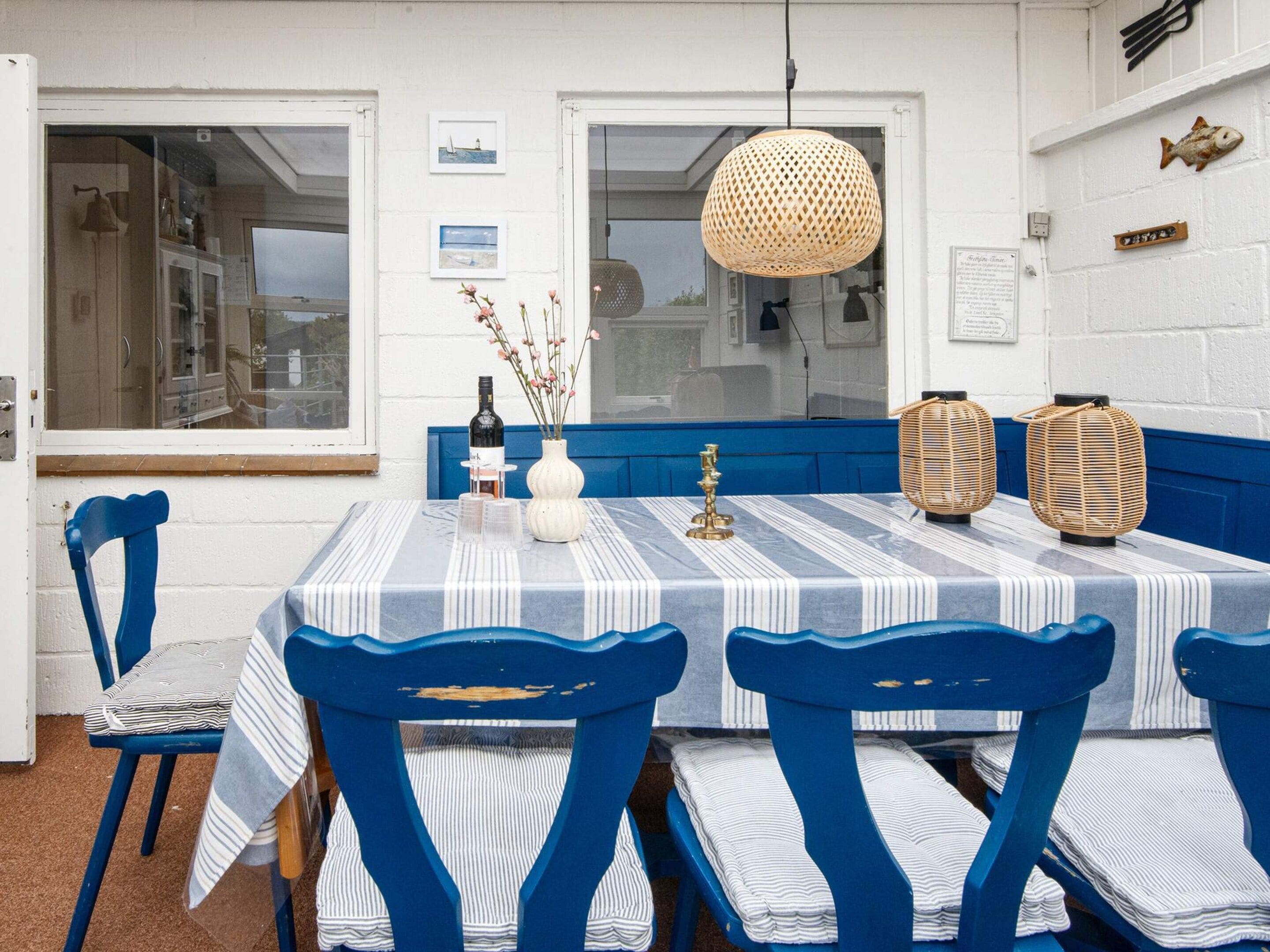 Een eethoek met een geruit tafelkleed, blauwe stoelen, een hanglamp en een raam met uitzicht op zee.