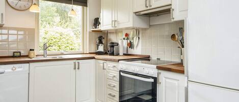 A kitchen with white cabinets, a black and white checkered floor, a clock on the wall, and a window with a view of greenery.