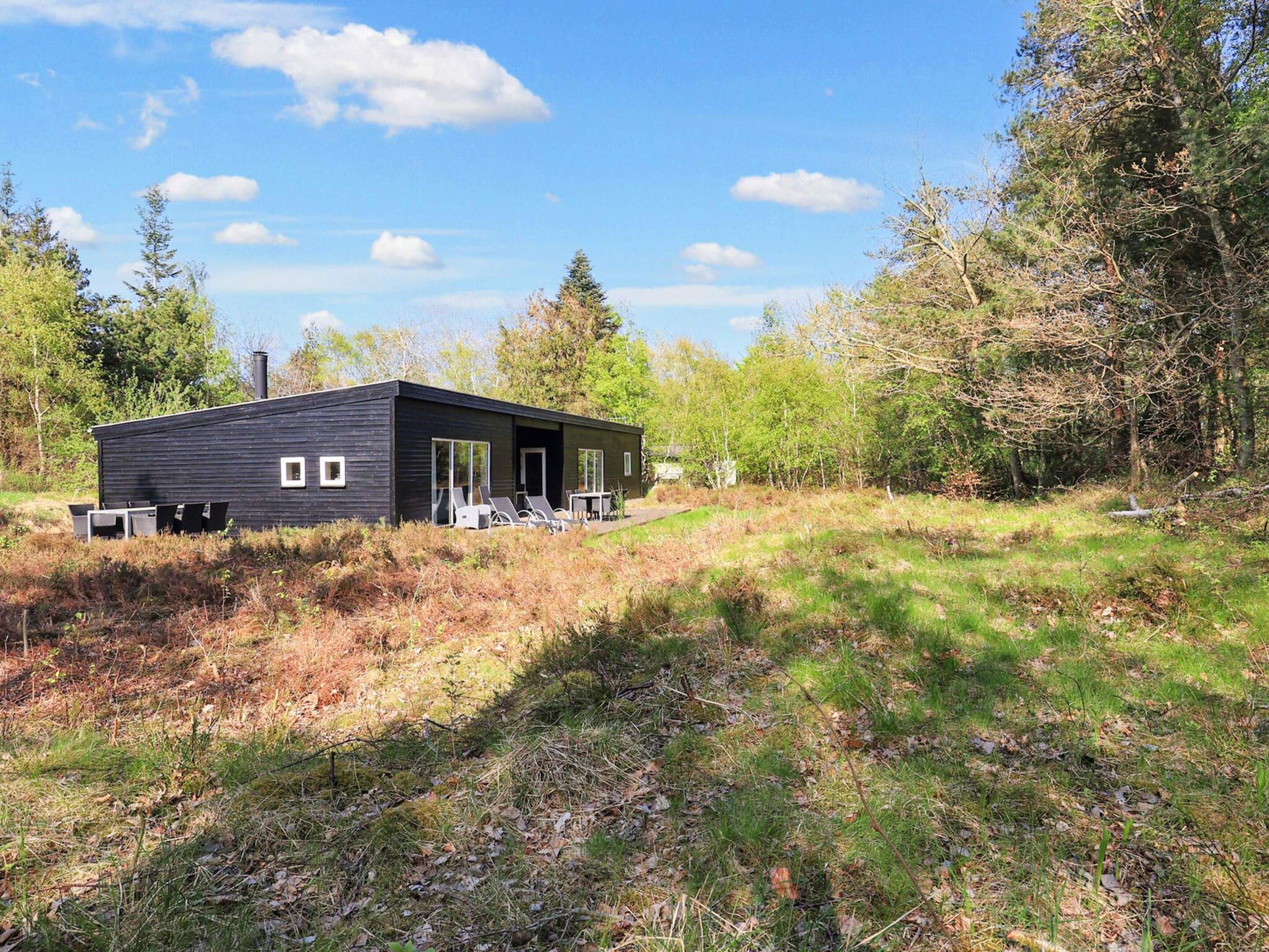 Une cabane moderne dotée d’un toit plat, de grandes fenêtres et d’une cheminée. Elle est entourée d’un paysage naturel, avec des arbres et de l’herbe.