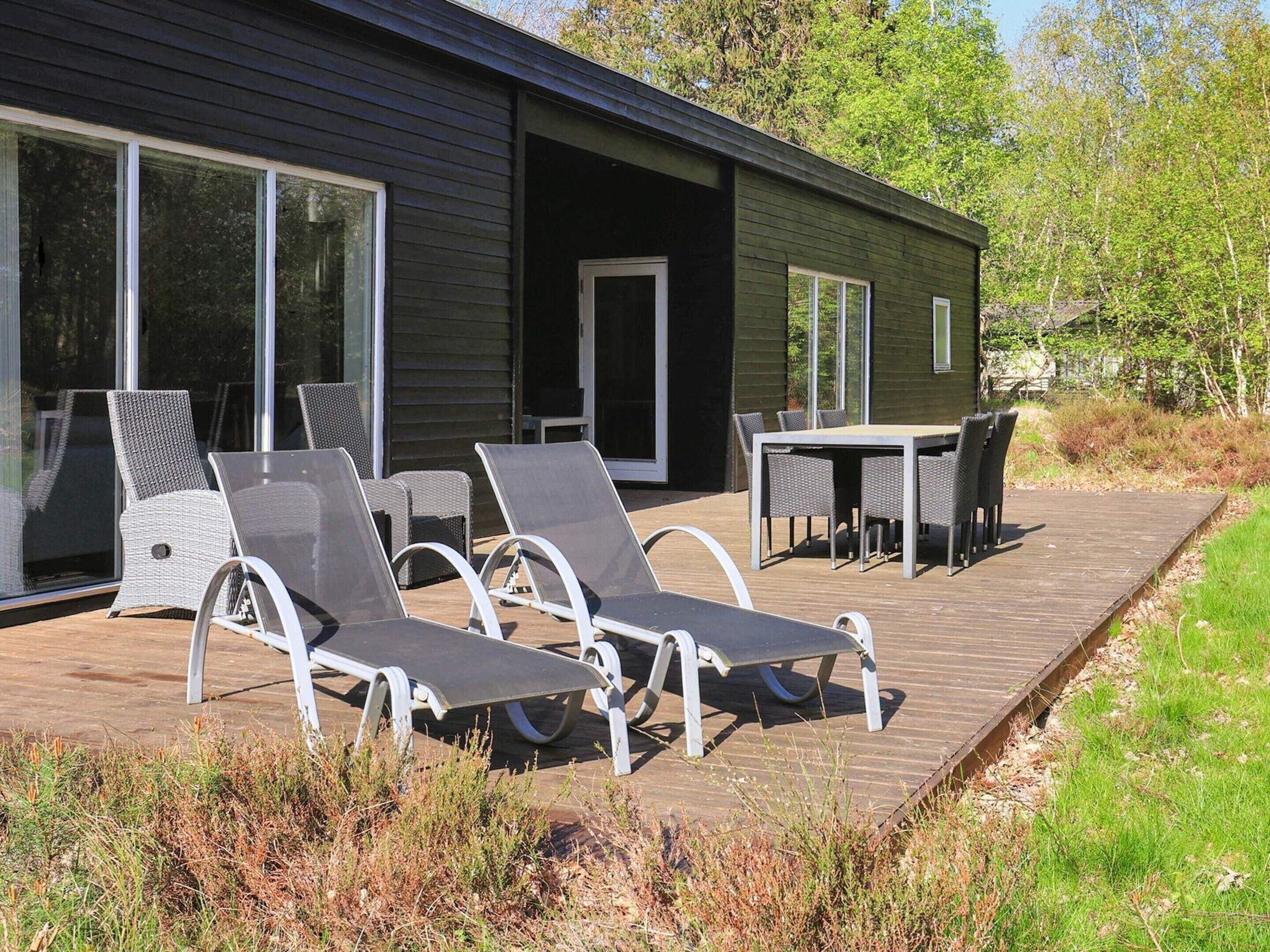 A modern outdoor deck with lounge chairs and a table, surrounded by a wooden deck and vegetation.