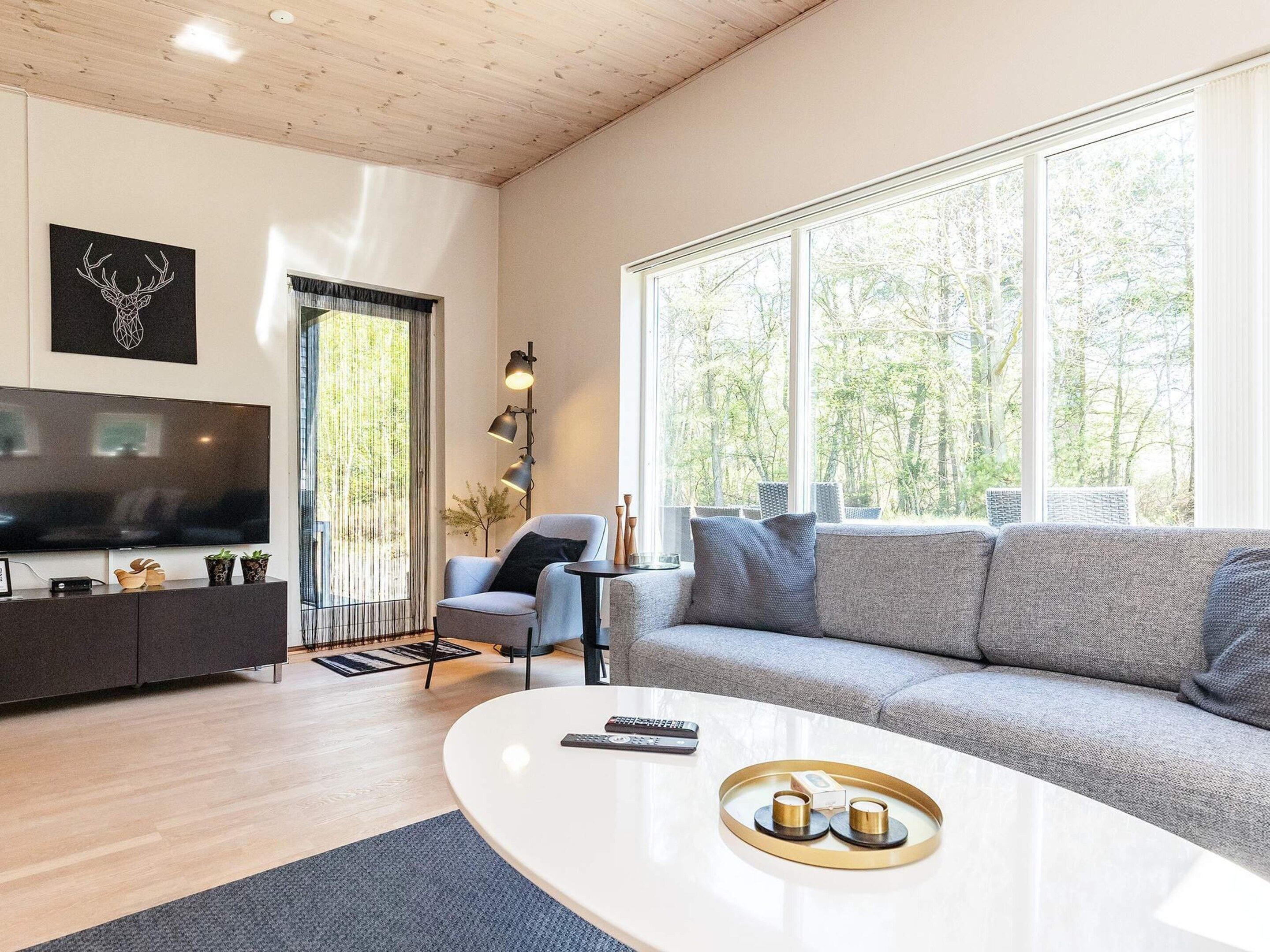 Ein modernes Wohnzimmer mit einem Flachbildfernseher, einem grauen Sofa, einem Couchtisch und einem großen Fenster mit Blick auf Bäume.