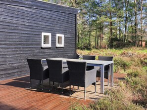 An outdoor patio with a table and chairs, a wooden deck, and a dark wooden wall with two windows. - 5 Star Holiday Home in Hadsund (Hadsund)