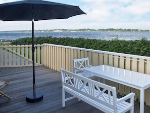 Um terraço com mesa e cadeiras brancas, um guarda-sol preto e vista para a água.
