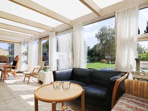 A sunlit room with a black leather sofa, a round wooden table, wicker chairs, and large windows with white curtains.