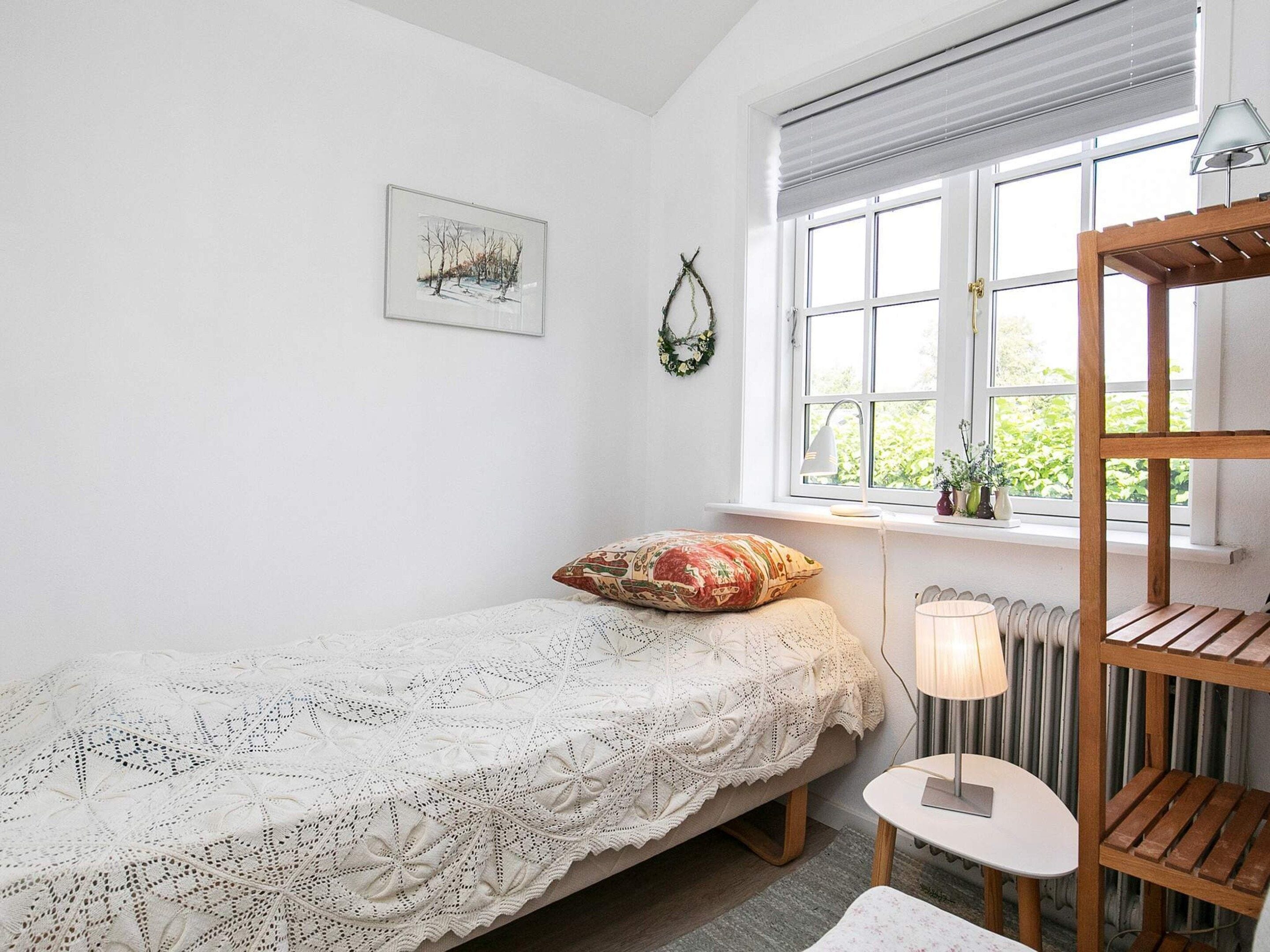A small, cozy bedroom with a single bed, a bedside table, a lamp, a radiator, and a window with a view of greenery.