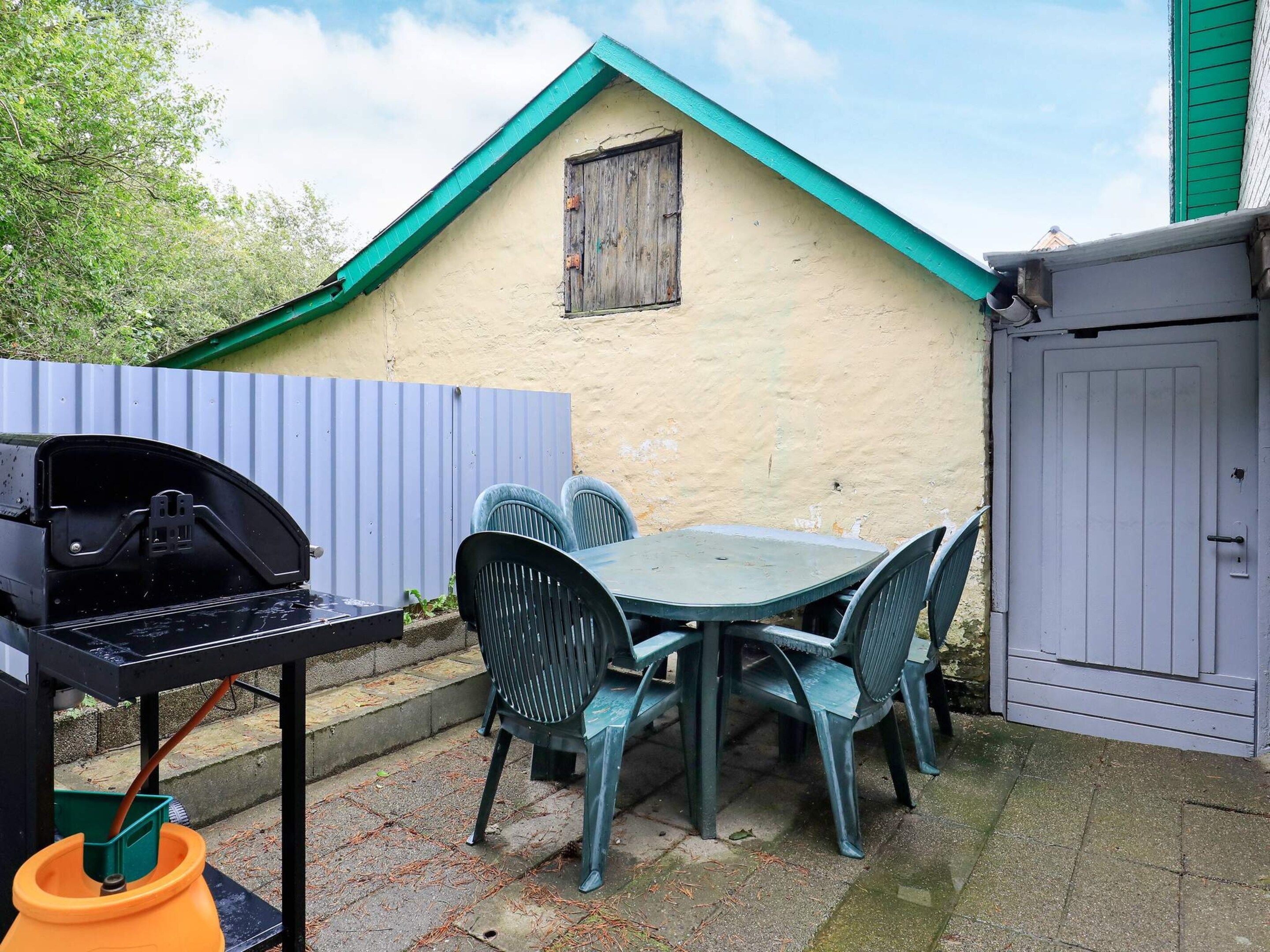 Une terrasse avec un barbecue, une table, des chaises et un petit bâtiment doté d’une fenêtre.