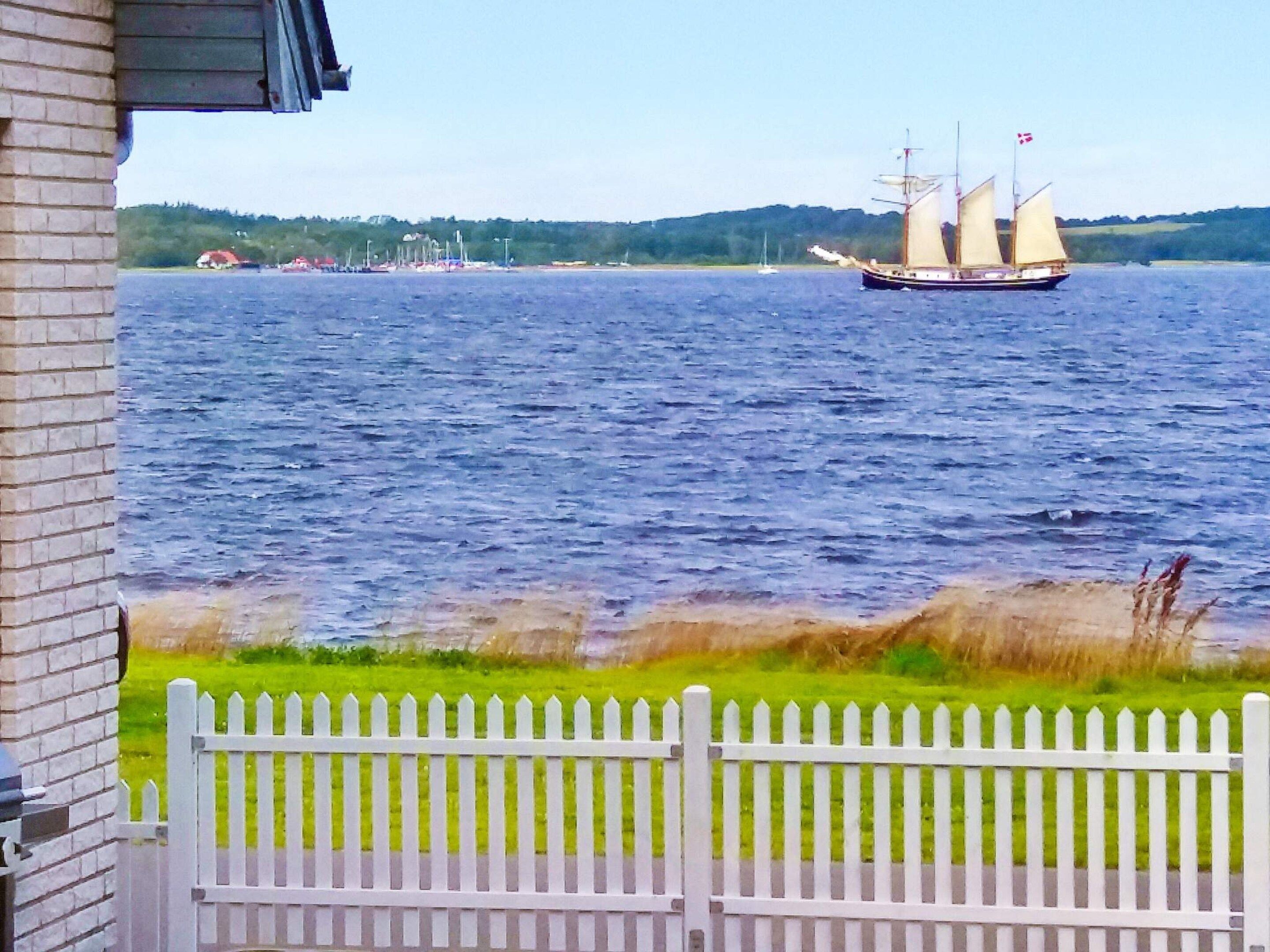 A waterfront view with a sailboat, a white picket fence, and a building with a brick facade.