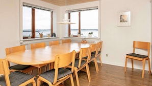 Een eetruimte met een grote houten tafel en stoelen, grote ramen met uitzicht op het water en een hanglamp.