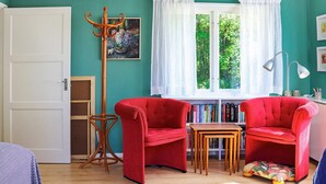 A room with two red armchairs, a wooden side table, a floor lamp, a bookshelf, and a window with a view of trees.