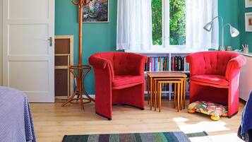 A room with two red armchairs, a wooden side table, a floor lamp, a bookshelf, and a window with a view of trees.