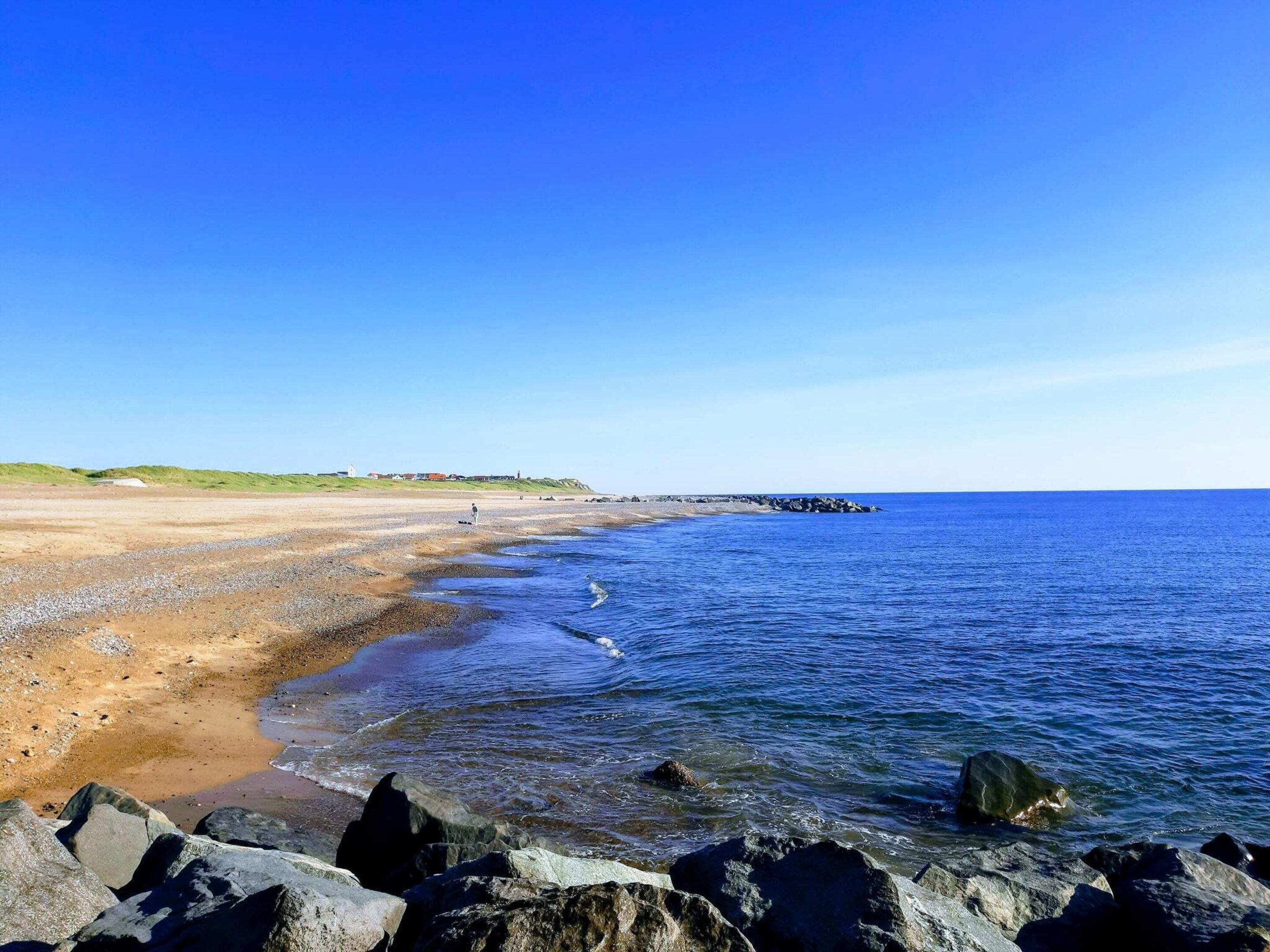 A coastal beach with clear skies, rocky shoreline, and distant buildings.