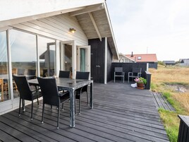 Una terraza de madera con una mesa y sillas, una planta en maceta y vista a un edificio con tejado inclinado.