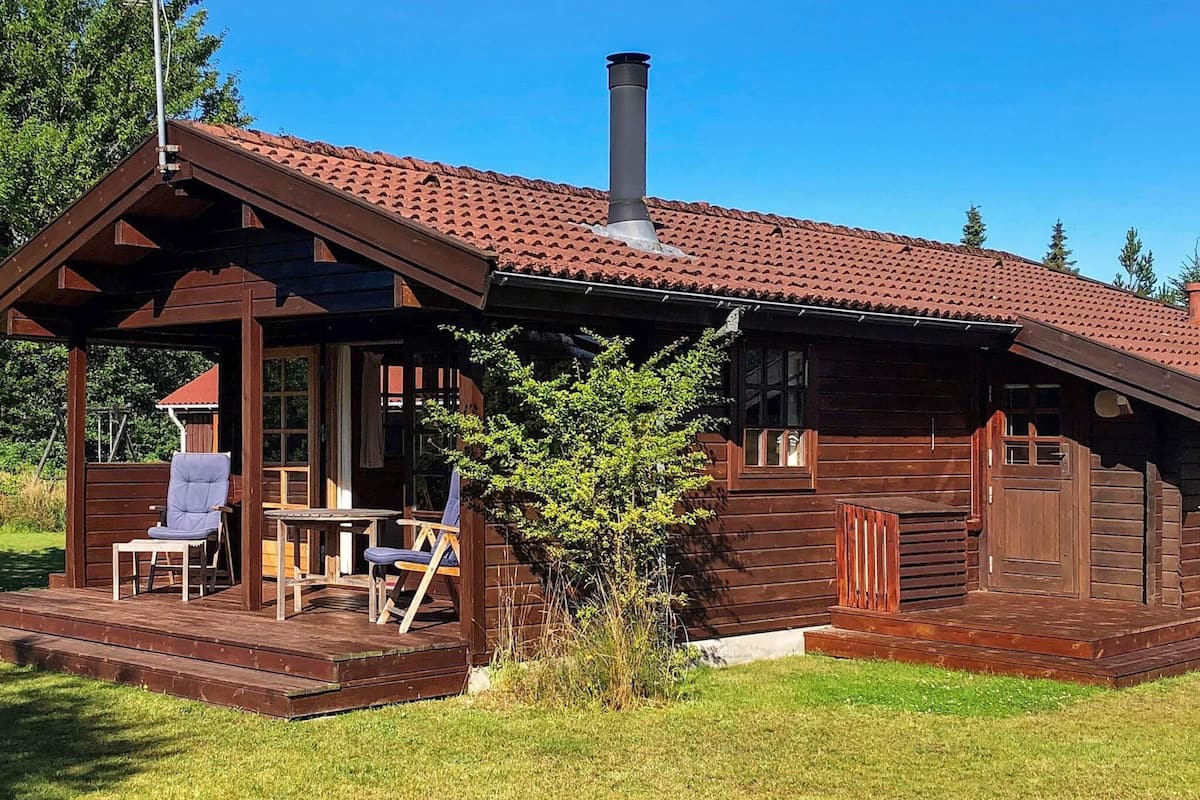 A wooden cabin with a tiled roof, a chimney, a porch with seating, and a small tree in front.