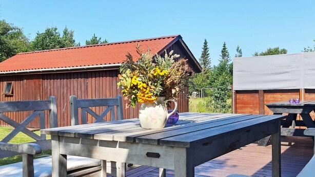 A wooden deck with a table and chairs, a vase with flowers, and a red-roofed building in the background. - Charming Coastal Retreat-by Traum (Jerup)