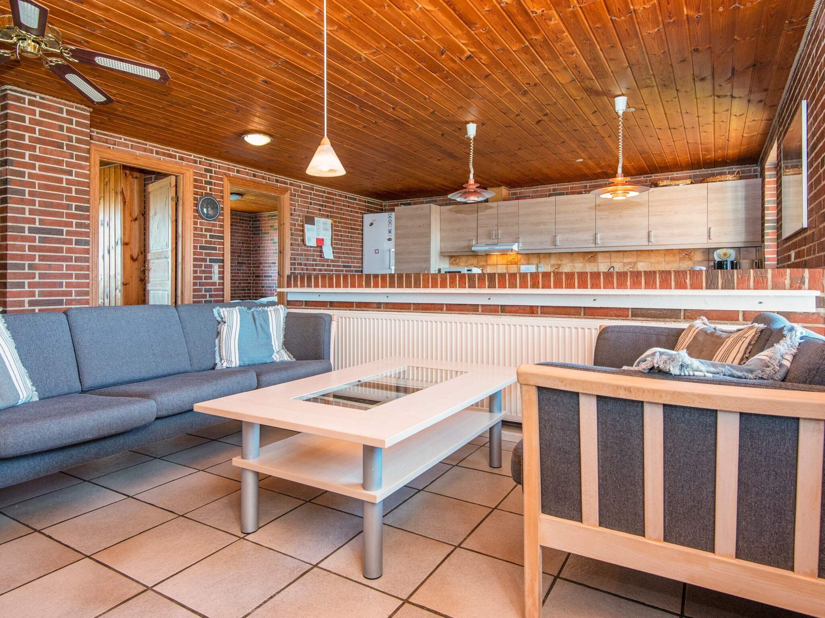 A cozy interior with a wooden ceiling, a brick wall, a sofa, a table, and a kitchen area.