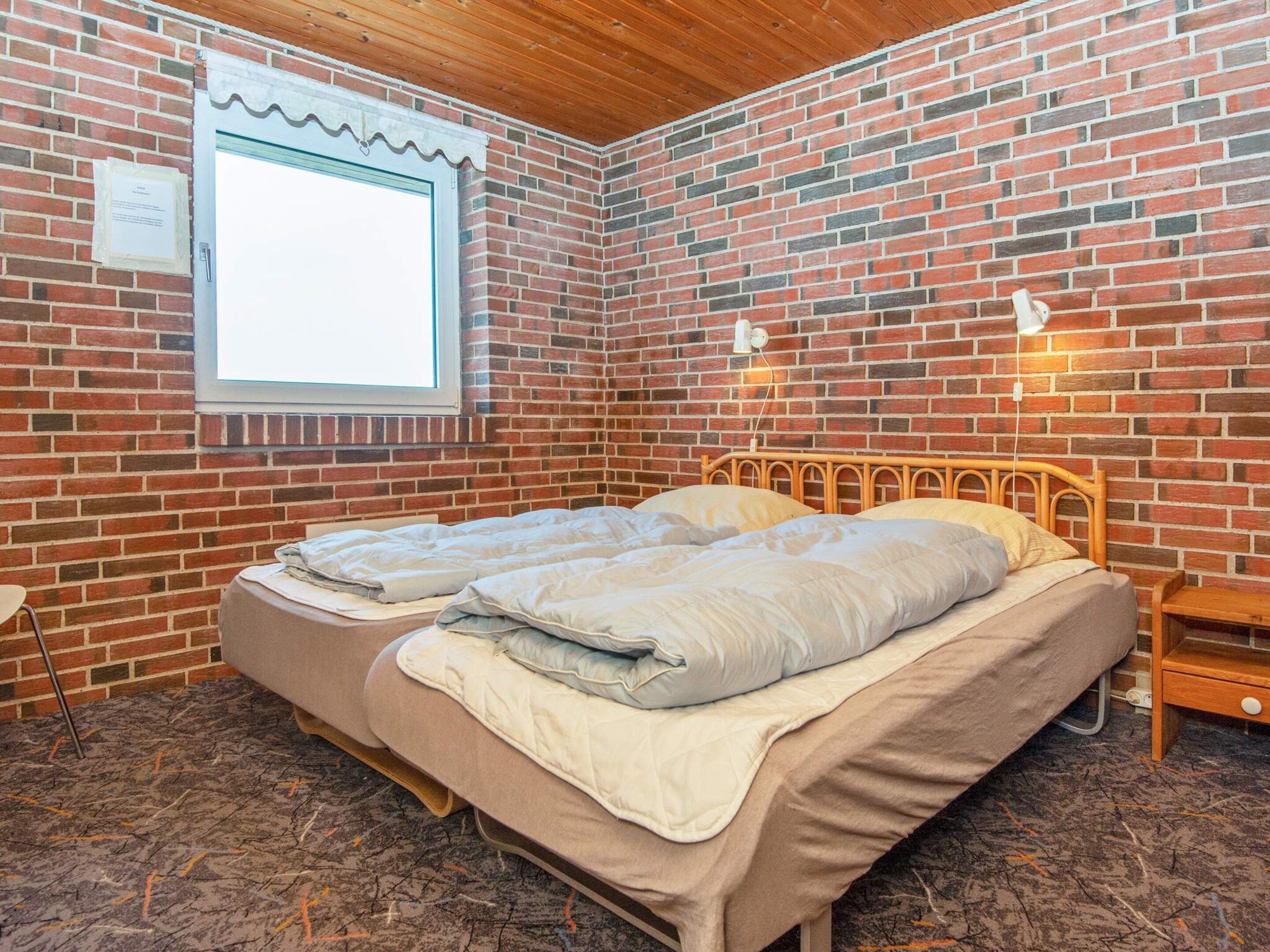 Une pièce avec un mur en briques, une fenêtre, un lit avec deux oreillers, une table de chevet et une lampe fixée au mur.