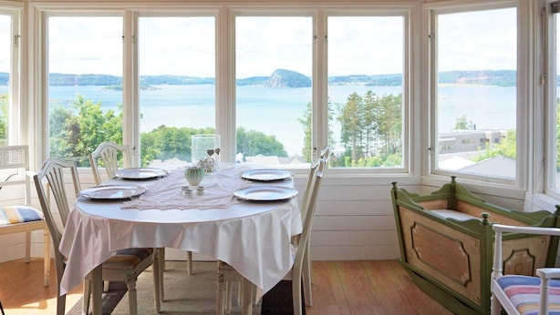 A dining area with a table set for four, large windows with a lake view, wooden ceiling beams, and a green upholstered bench. - 5 Person Holiday Home in Uddevalla (Uddevalla)