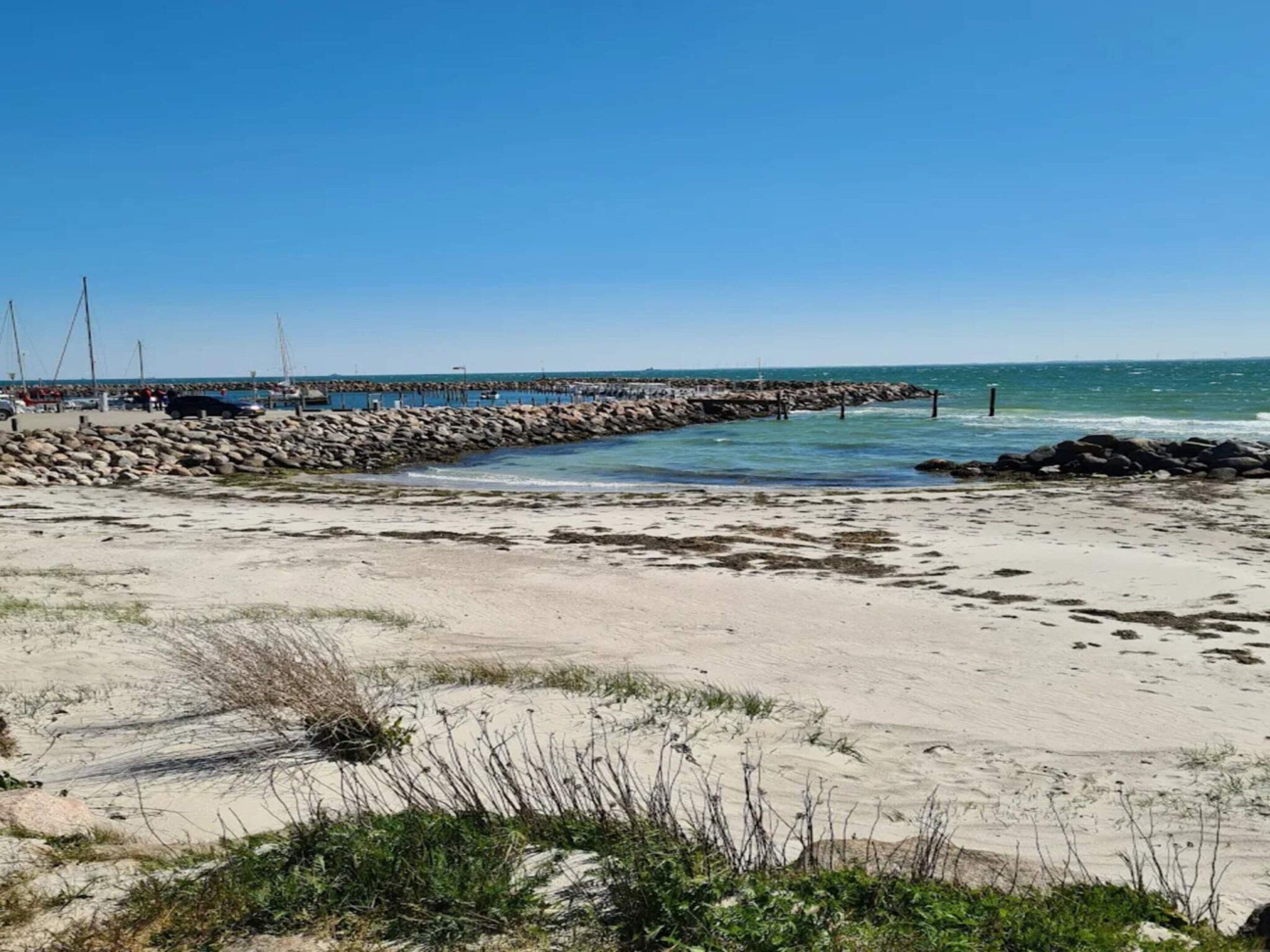 A sandy beach with a rocky breakwater extending into the water, clear skies, and sparse vegetation.