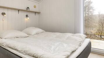 Een bed met grijs frame en wit beddengoed, een houten plank met een plant en een rond voorwerp, en een raam met uitzicht op bomen.