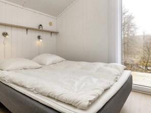 En säng med en grå ram och vita sängkläder, en trähylla med en växt och ett runt föremål, och ett fönster med utsikt över träd.