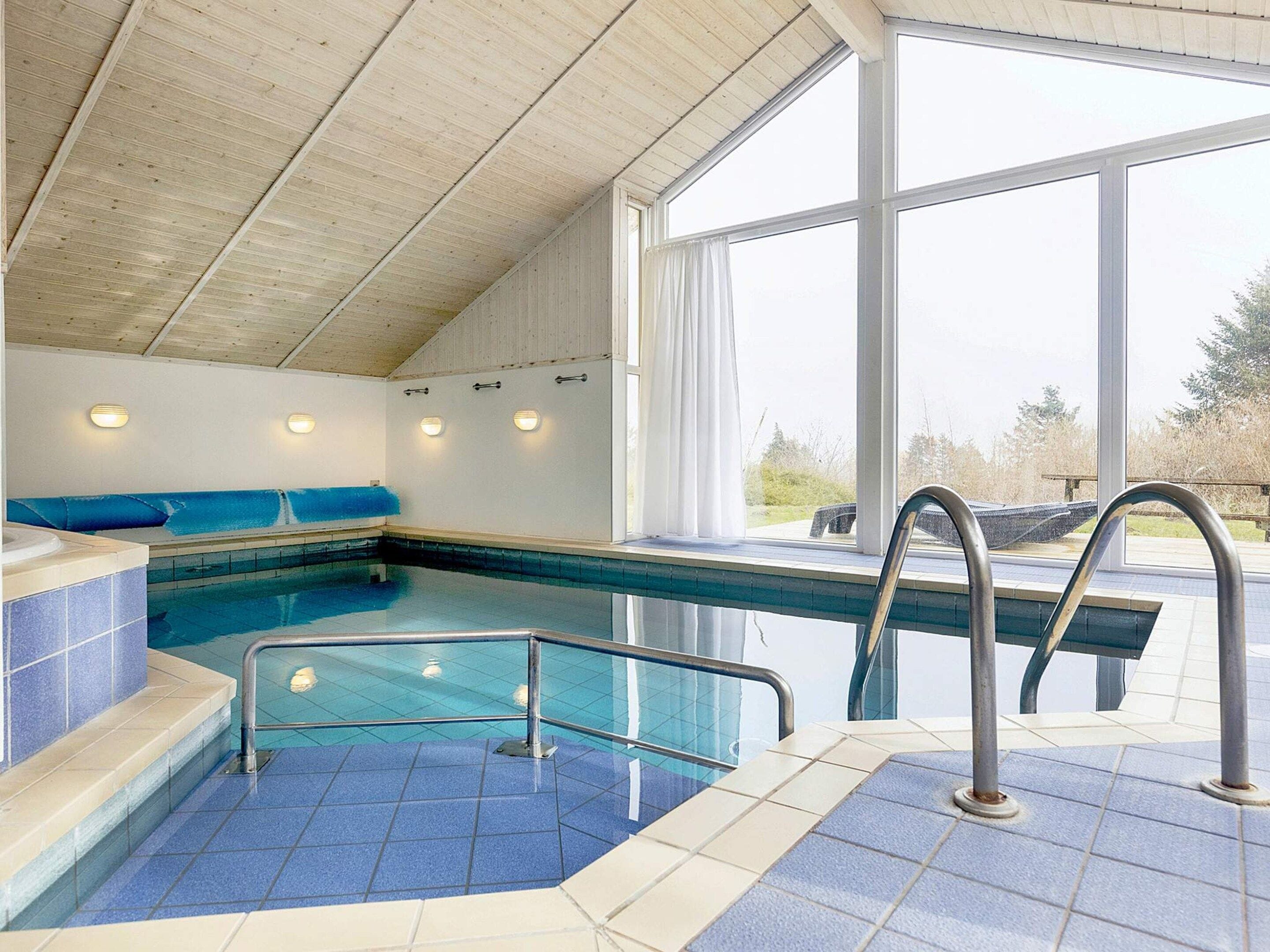 An indoor swimming pool with a clear view of the outdoors, a blue-tiled floor, and a bench along the wall.
