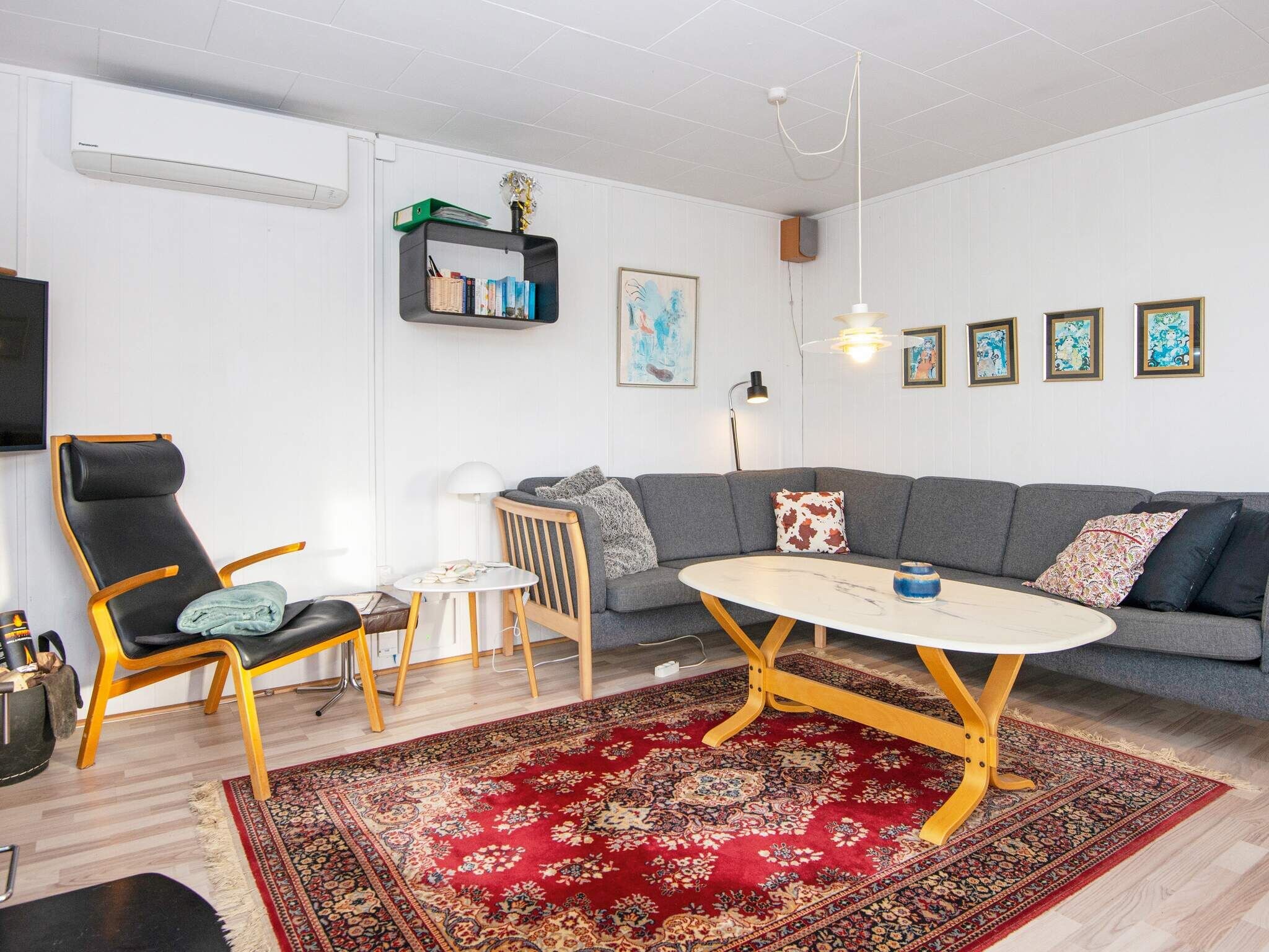 Una sala moderna con un sofá gris, una mesa redonda de centro, un tapete rojo estampado, un sillón de cuero negro y una pequeña mesa blanca con una lámpara.
