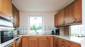 Una cocina moderna con armarios de madera, horno empotrado, microondas, cafetera y un amplio ventanal con vistas a la vegetación.