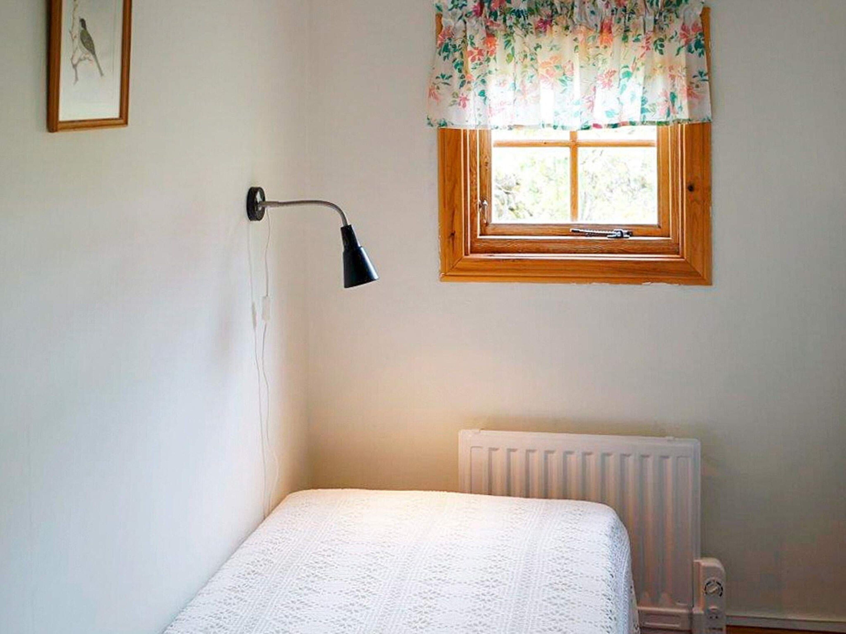 A bedroom with a single bed, a window with a floral curtain, a wall-mounted lamp, and a radiator.