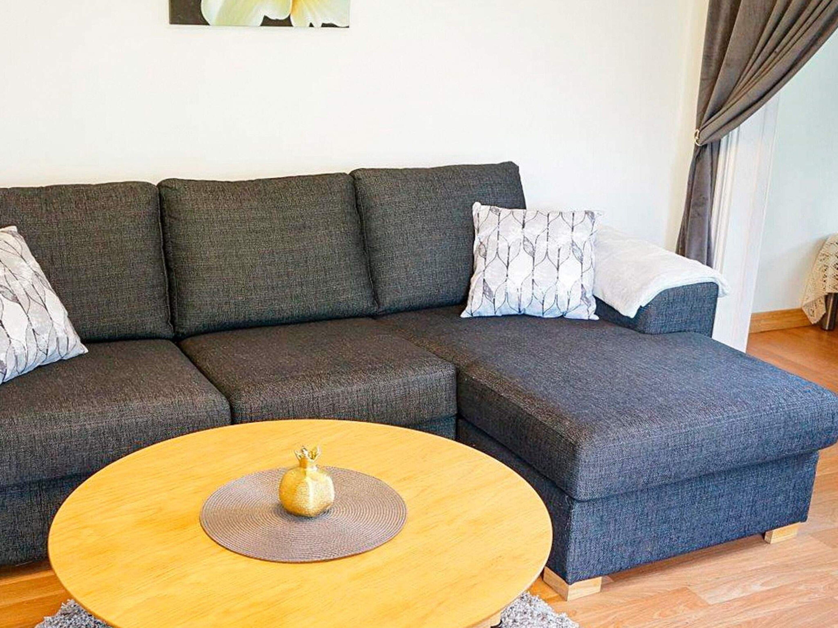 A sectional sofa with patterned cushions, a round wooden coffee table with a decorative item, and a plain wall with a single framed picture.