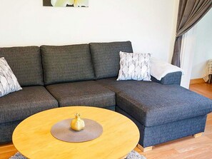 Un canapé modulable avec des coussins à motifs, une table basse ronde en bois agrémentée d’un objet décoratif, et un mur sobre orné d’un seul tableau encadré.
