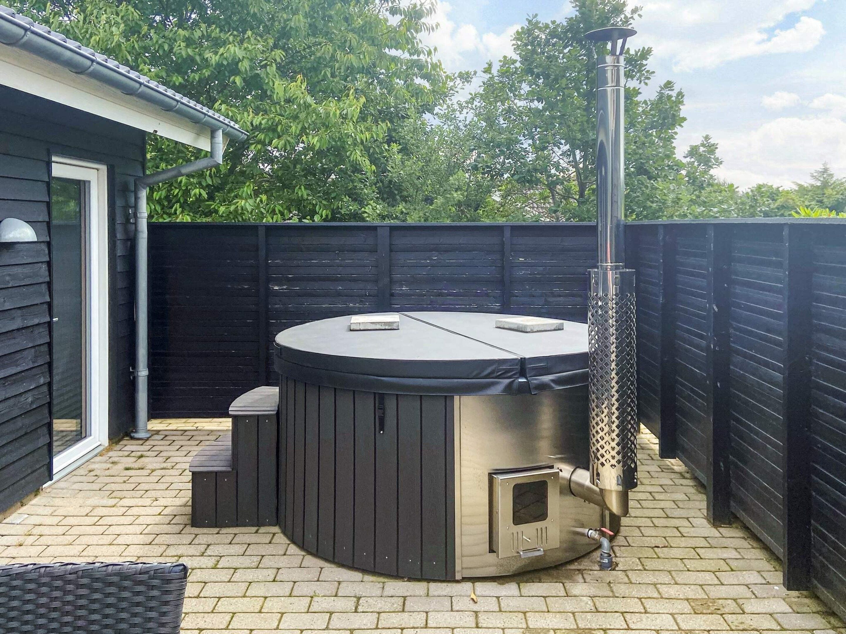 Un jacuzzi con chimenea, una mesita y una silla en un patio trasero con cerca negra y una casa al fondo.
