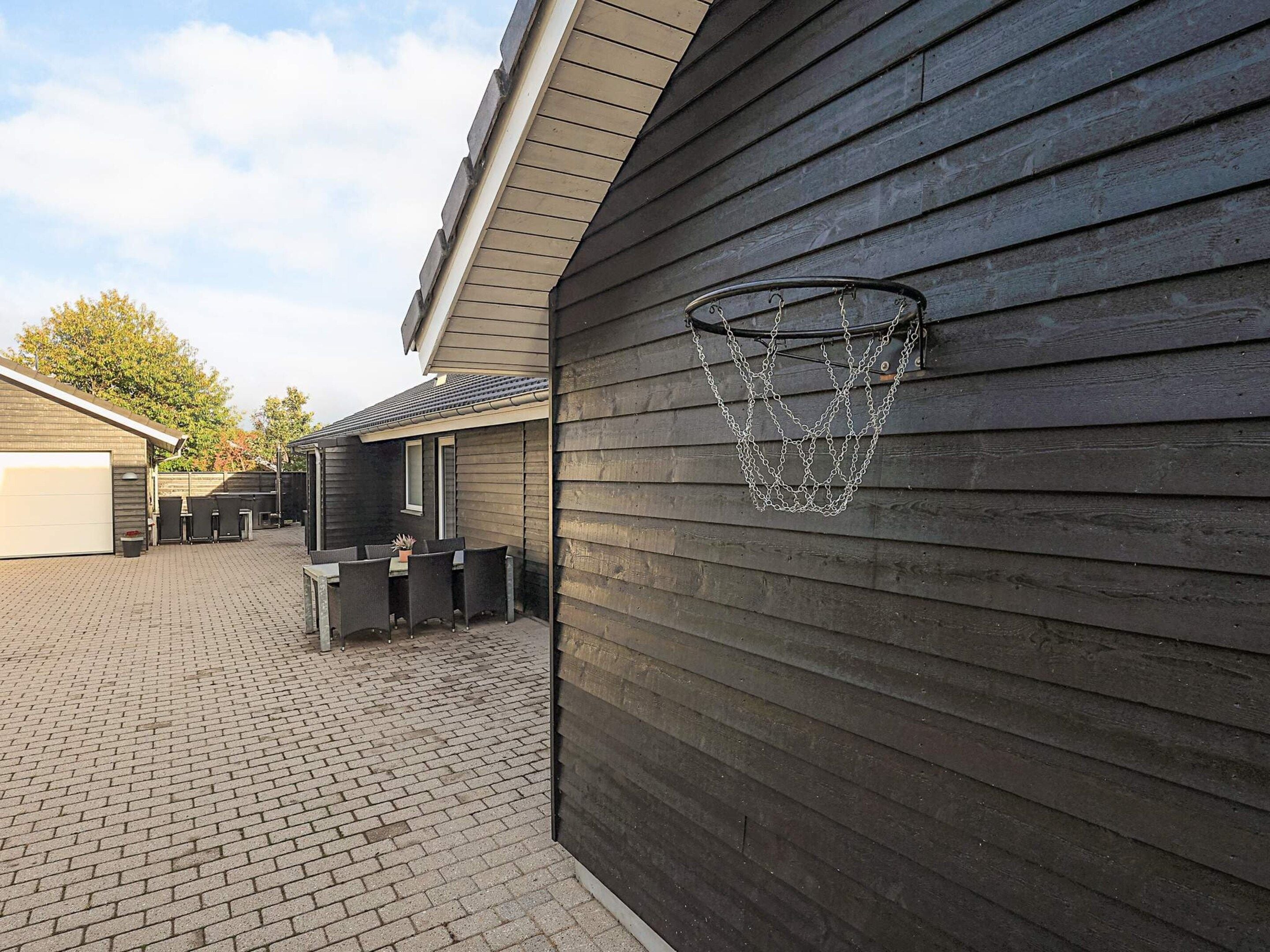 En basketkorg som är fastsatt på en vägg av trä, en uteplats med tegellagning och en byggnad med ett garage.