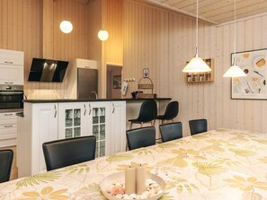 A modern kitchen with white cabinets, a built-in oven, and a refrigerator. There are two pendant lights above the counter and a dining table with chairs. - 5 Star Holiday Home in Hojslev (Hojslev)