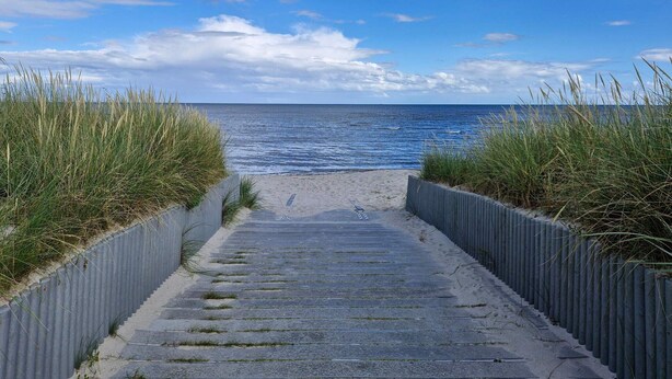 Un camino de concreto lleva a una playa, con vista despejada al océano y un cielo parcialmente nublado.