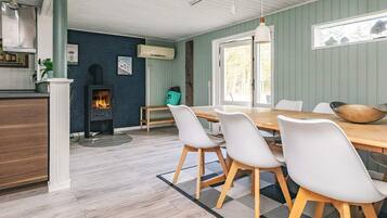 Et spiseområde med et træbord og hvide stole, en pejs og udsigt til naturen.