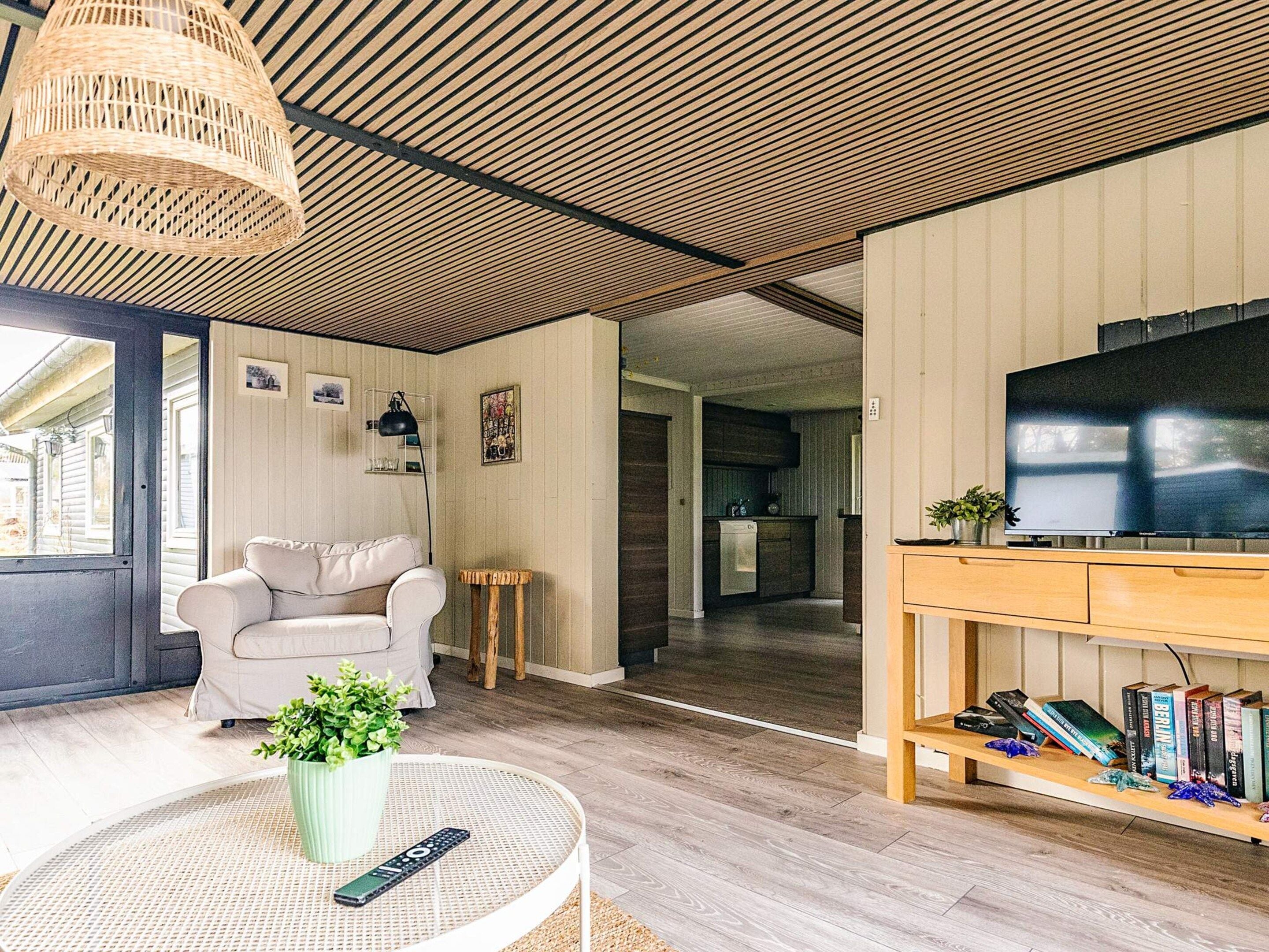 En stue med sofa, rundt bord med plante og fjernbetjening, TV på træstel og et køkken i baggrunden.