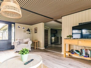 Ein Wohnzimmer mit einer Couch, einem runden Tisch mit Pflanze und Fernbedienung, einem Fernseher auf einem Holzständer und einer Küche im Hintergrund.