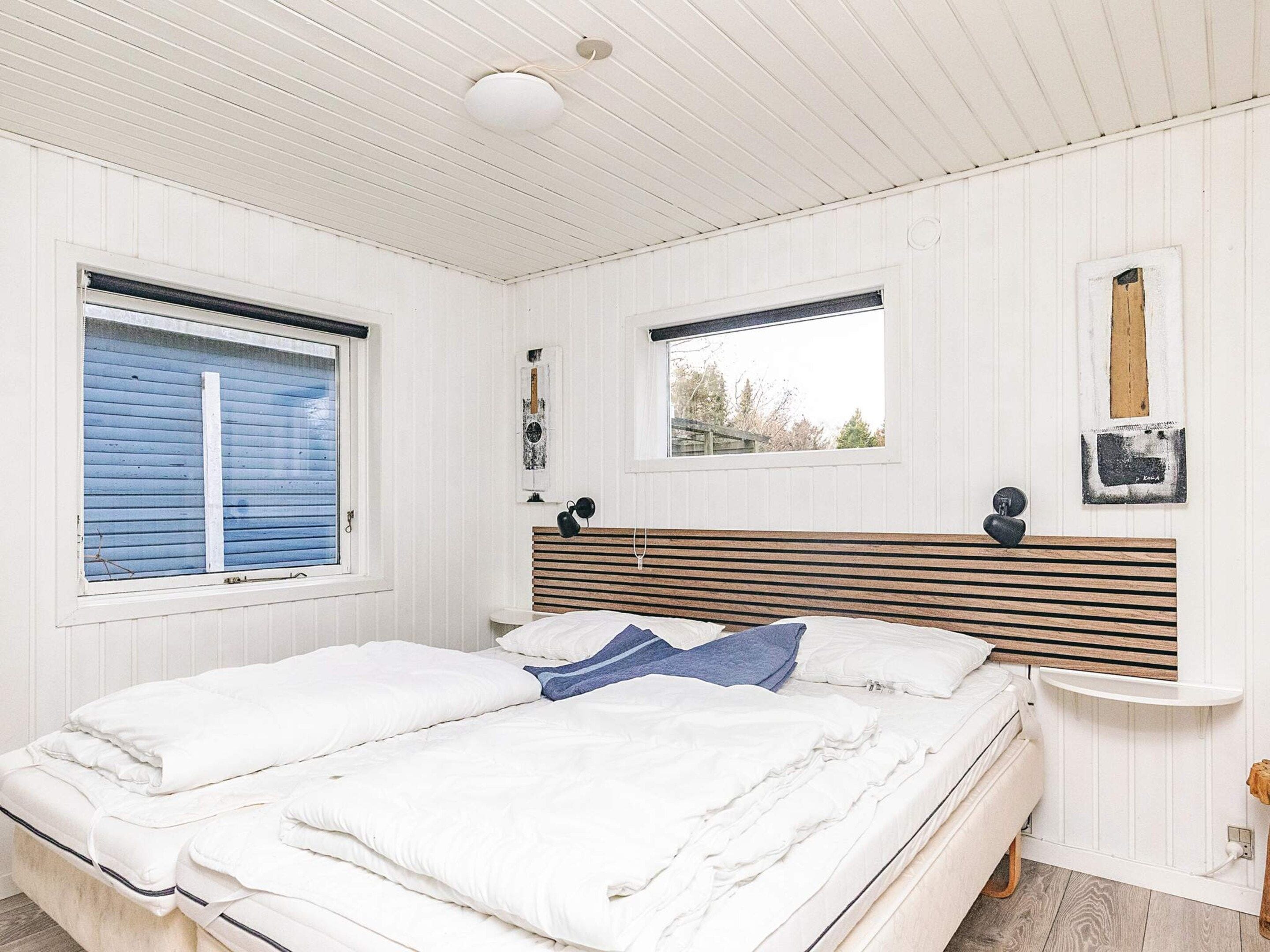 A bedroom with a bed, two windows, a bedside table, and a wall-mounted light fixture.