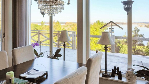 Un espace repas avec vue sur un lac et des arbres, un lustre et une terrasse en bois à l’extérieur.