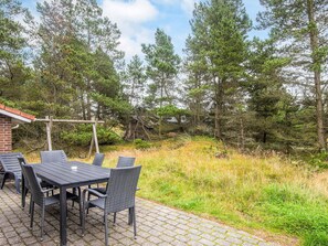 Une terrasse extérieure avec une table et des chaises, entourée d’un paysage naturel composé d’arbres et d’un bâtiment en briques en arrière-plan.
